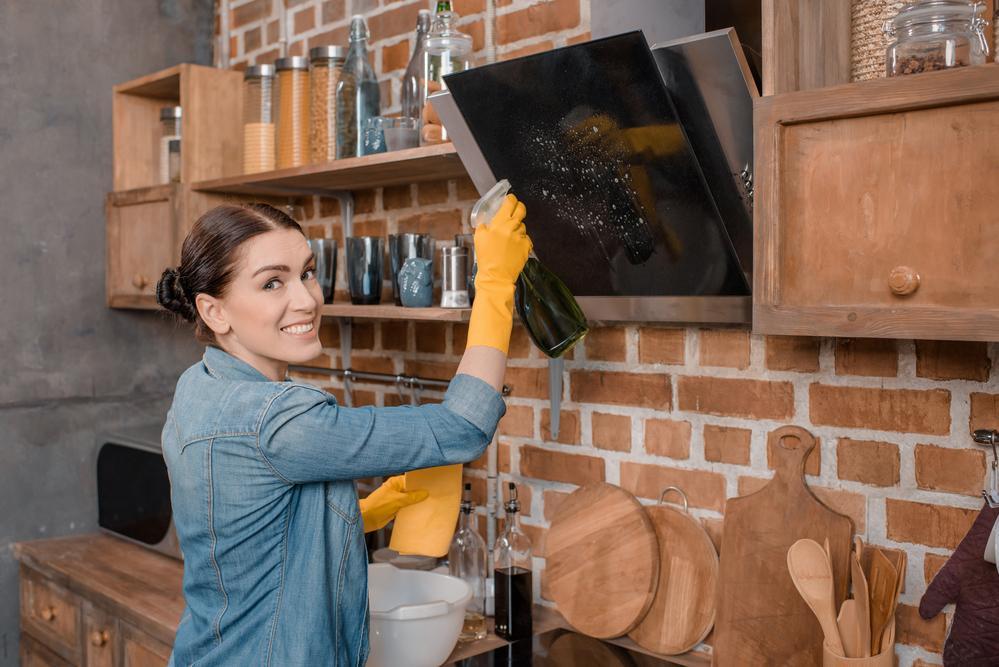 A woman cleaning the kitchen countertops and cabinets with a spray cleaner.  